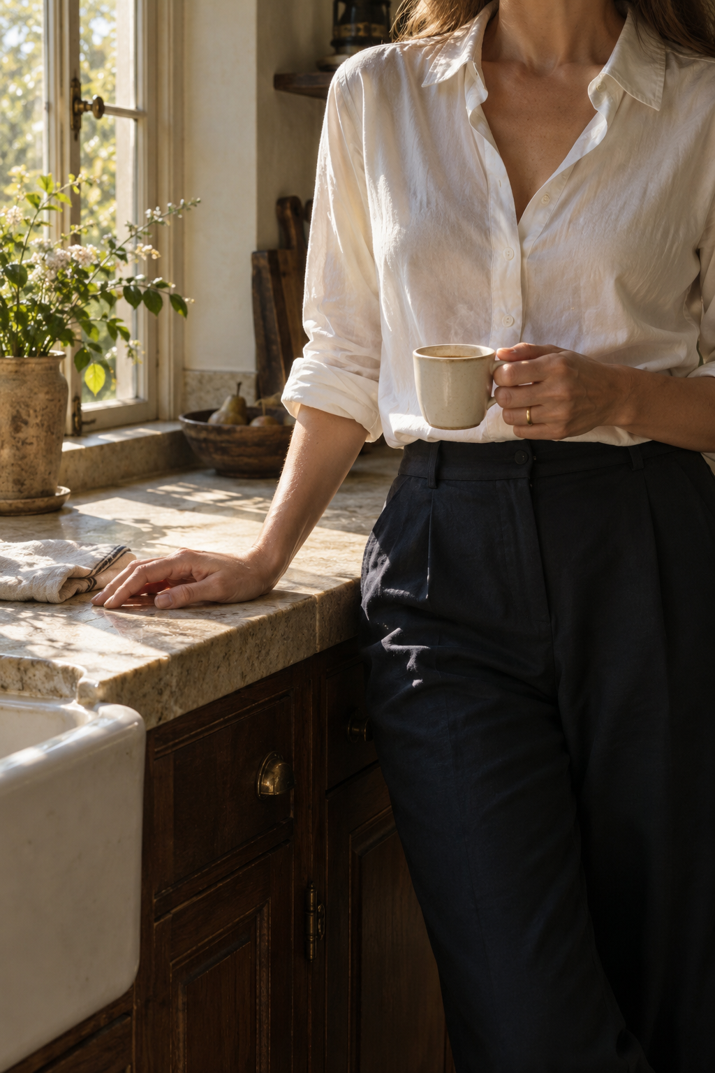 A woman holding her morning coffee beside a sunlit kitchen window, her face outside the frame